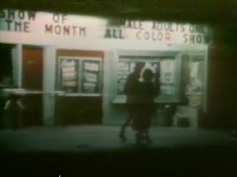 Two people walk in front of a theater at night with illuminated marquees reading "SHOW OF THE MONTH" and "ALL COLOR SHOW." Posters are displayed in glass cases by the entrance.