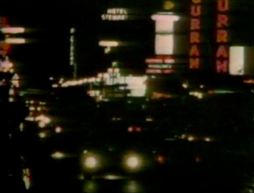 A busy city street at night with illuminated signs, including one reading "CURRAN," and headlights of several cars visible.
