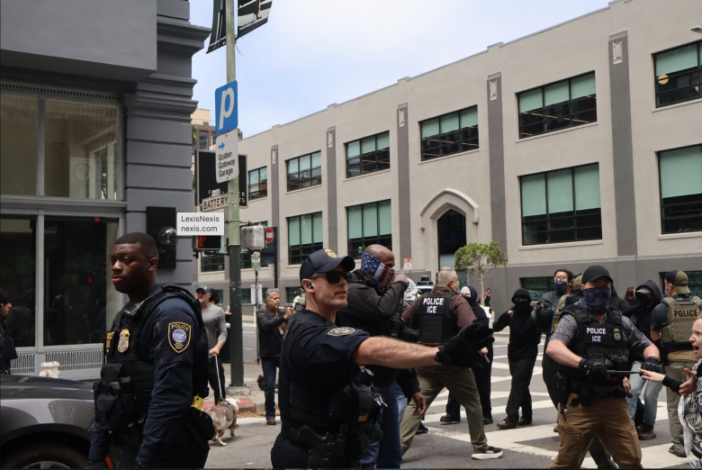 Police officers stand on a city street, some directing or holding back people, with a crowd and multi-story buildings visible in the background.