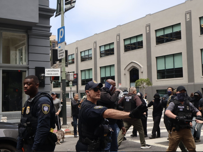 Police officers stand on a city street, some directing or holding back people, with a crowd and multi-story buildings visible in the background.