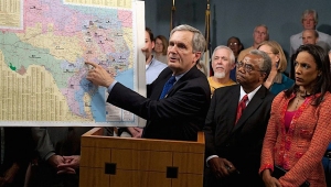 A man in a suit points to a map of Texas on an easel while speaking at a podium, surrounded by a group of people.