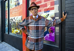 An older man wearing a brown hat and plaid jacket stands with one hand on a black wall beside a window with colorful posters and graffiti.