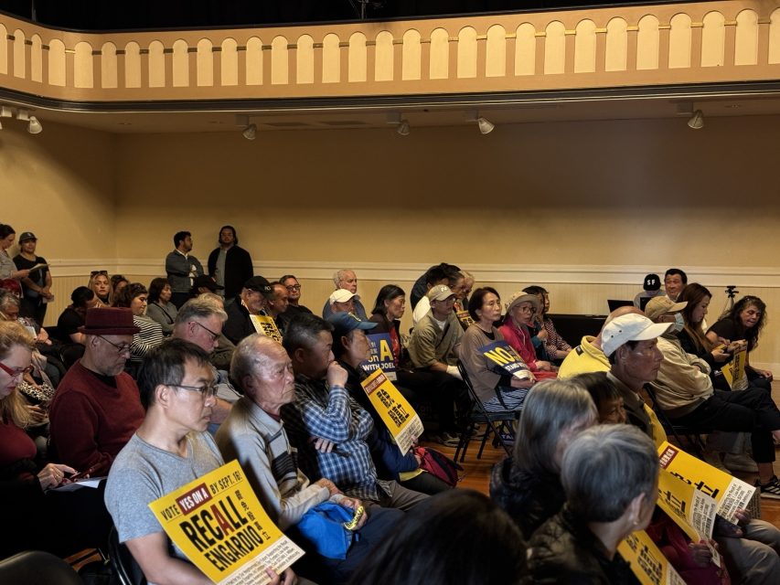 A group of people sit in a meeting room, many holding yellow protest signs that read “RECALL ENGARDIO”.