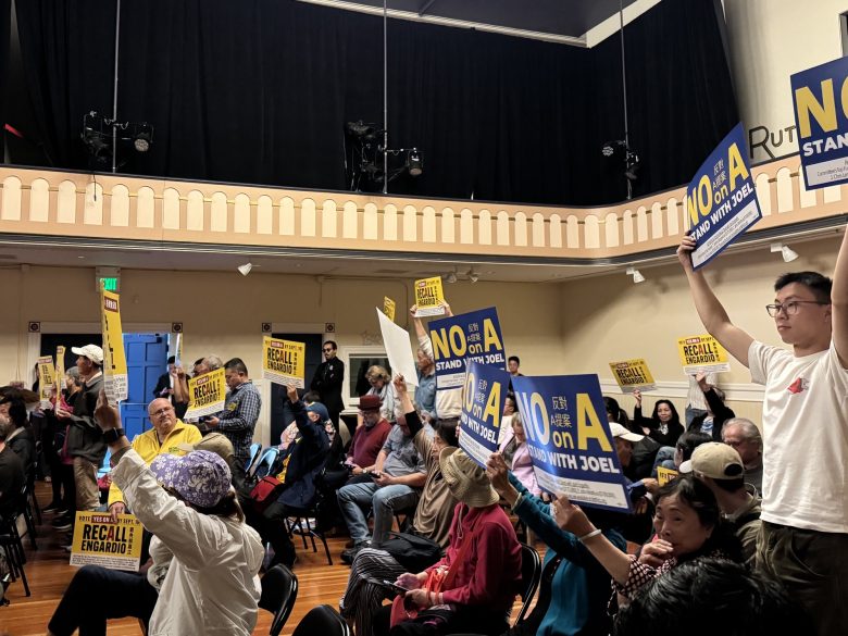 People seated and standing in a meeting room hold up signs that read "NO on A" and "Recall Engardio," indicating opposition to a recall measure.