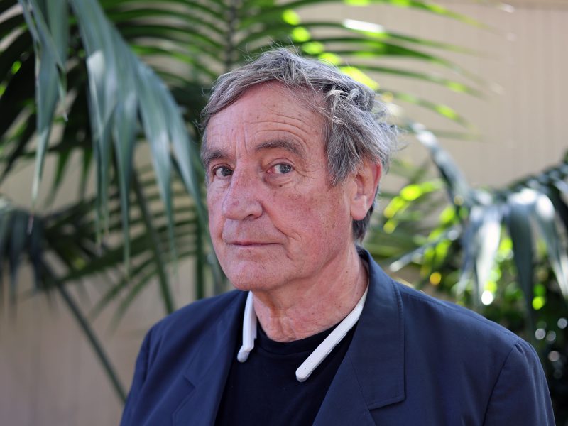 An older man with gray hair stands in front of large green plants, wearing a dark jacket and a black shirt, looking slightly to the side.