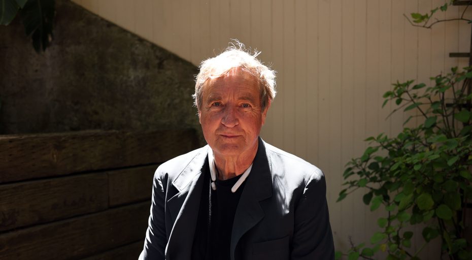 An older man with light hair wearing a dark blazer sits outdoors in sunlight, with a wooden wall and greenery in the background.