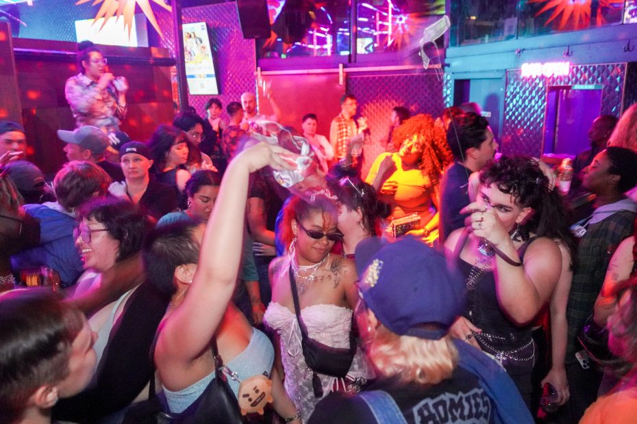 A crowded nightclub scene with people dancing, talking, and holding drinks under colorful lights.