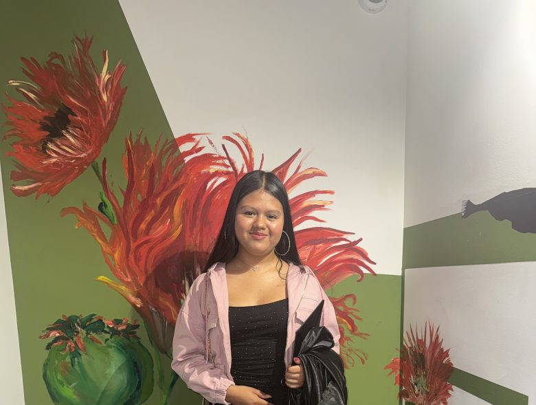 A young woman stands in front of a wall with large painted red flowers, wearing a light pink jacket over a black top and holding a black coat.