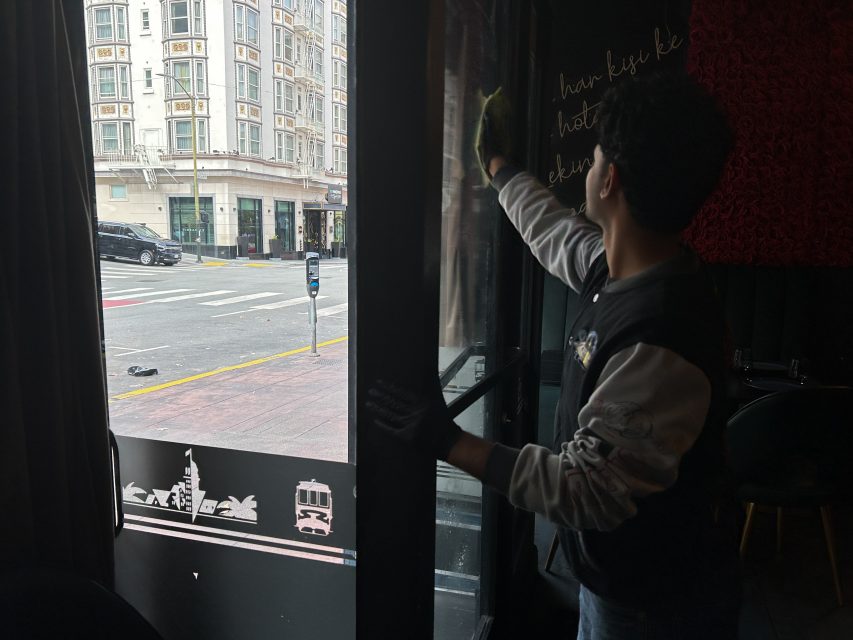 A person wearing gloves cleans the inside of a glass door at a restaurant, with a city street and buildings visible outside through the door.