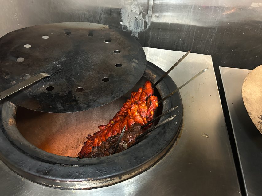 Several skewers of marinated meat are being cooked inside a traditional tandoor oven, partially covered by a metal lid, in a restaurant kitchen setting.