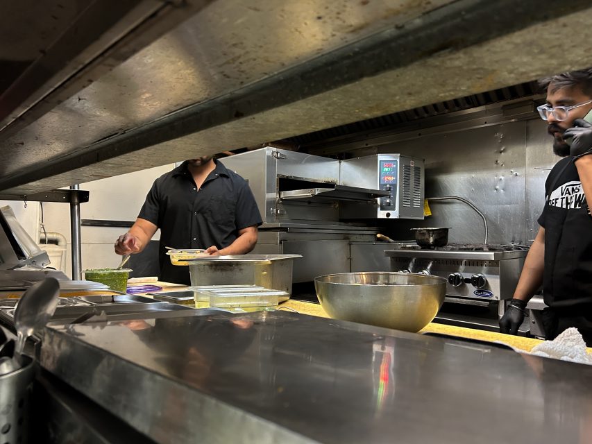 Two people work in a commercial kitchen, preparing food at a counter with various containers, a metal bowl, and cooking equipment visible in the background.