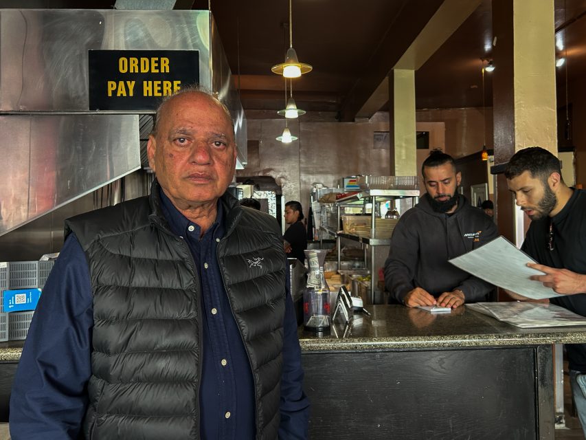 A man in a black vest stands in front of a counter at a restaurant, with two staff behind him looking at papers. A sign above reads "ORDER PAY HERE.