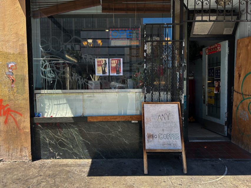 Street view of a restaurant entrance with a chalkboard sign advertising “ANY Coffee $1.50”, flyers on the window, and graffiti on the exterior wall and glass.