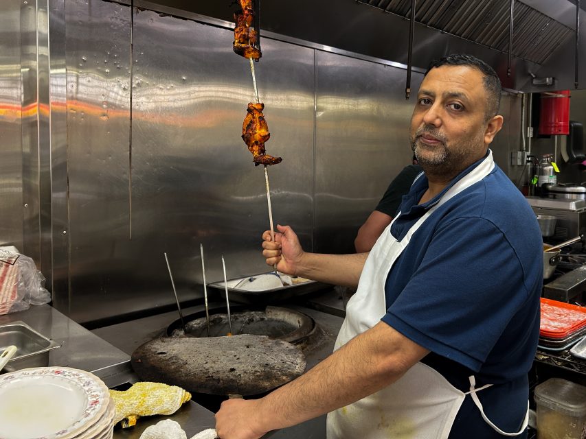 A man wearing an apron holds a skewer with grilled meat over a tandoor oven in a commercial kitchen.