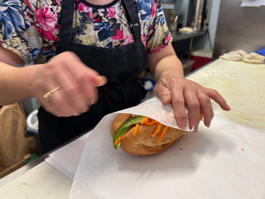 A person in a floral shirt and black apron wraps a sandwich with vegetables in white paper on a kitchen counter.
