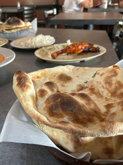 A basket of naan bread in the foreground, with plates of rice and grilled chicken on a restaurant table in the background.