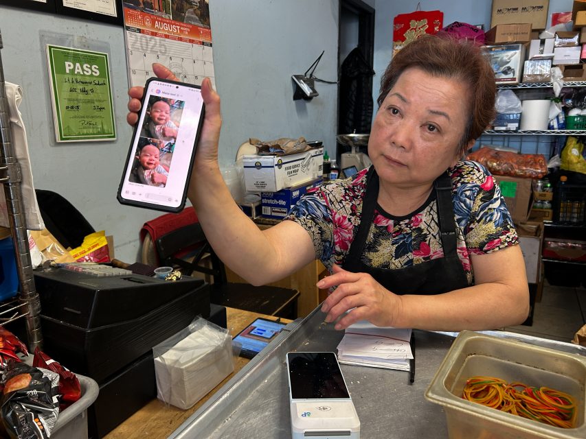 A woman behind a counter holds up a smartphone displaying photos of a baby, with various supplies and items visible in the background.