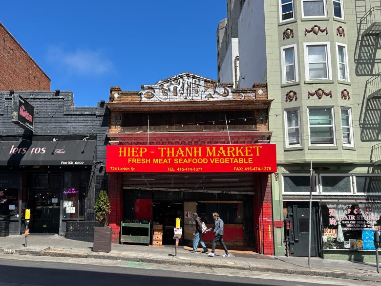 Hiep-Thanh Market storefront with a red sign, located between other businesses on a sunny street; two people walk past the entrance.