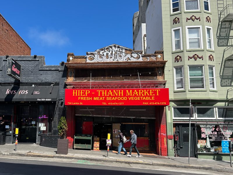 Hiep-Thanh Market storefront with a red sign, located between other businesses on a sunny street; two people walk past the entrance.