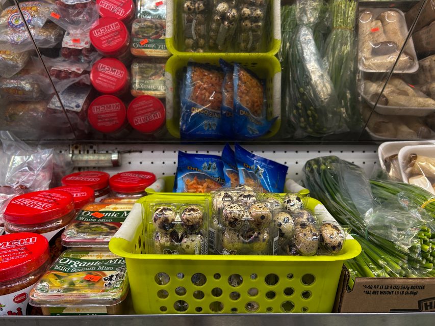 A yellow basket of quail eggs sits in a refrigerated display with tofu, packaged vegetables, greens, and containers of organic miso and fish roe.