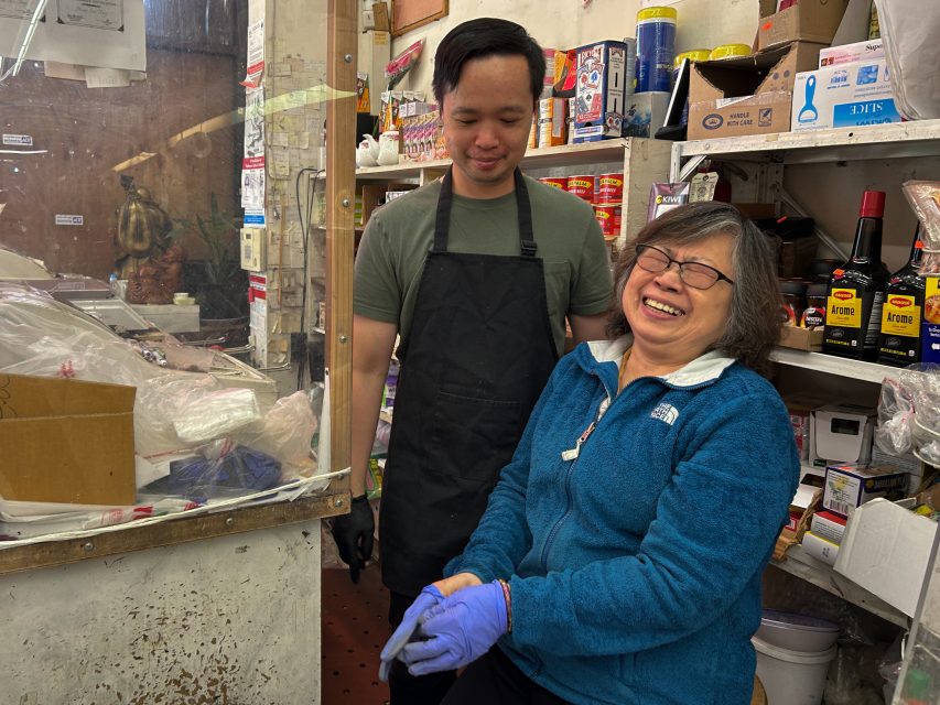 A smiling woman sits wearing gloves and a blue jacket, while a man in a black apron stands beside her in a cluttered store or kitchen.