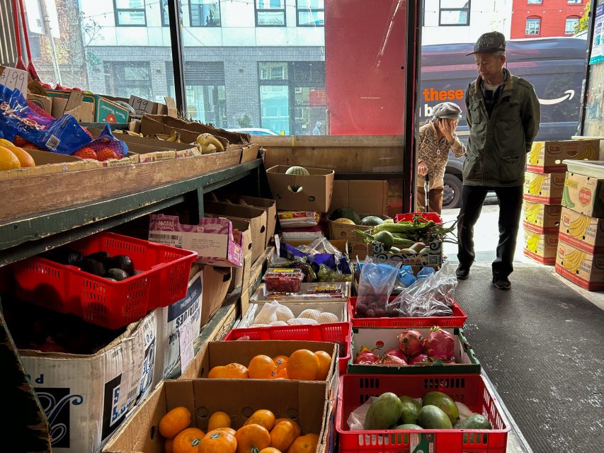 A man stands inside a market near boxes of assorted fruits and vegetables; another person walks outside near the entrance.