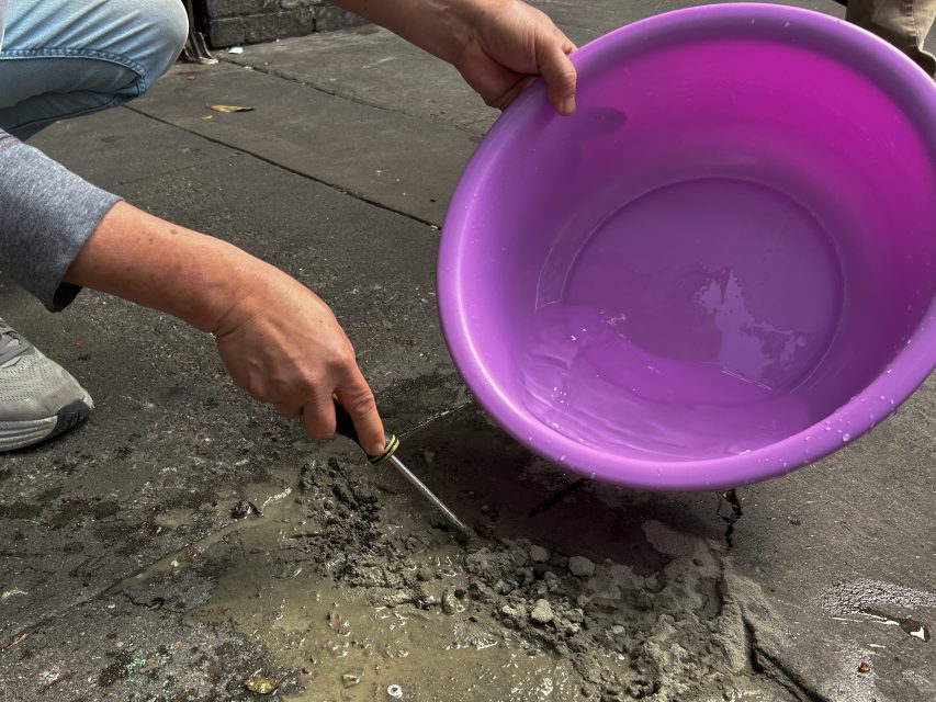 A person uses a knife to mix wet cement on pavement, holding a large purple plastic basin partially filled with water.