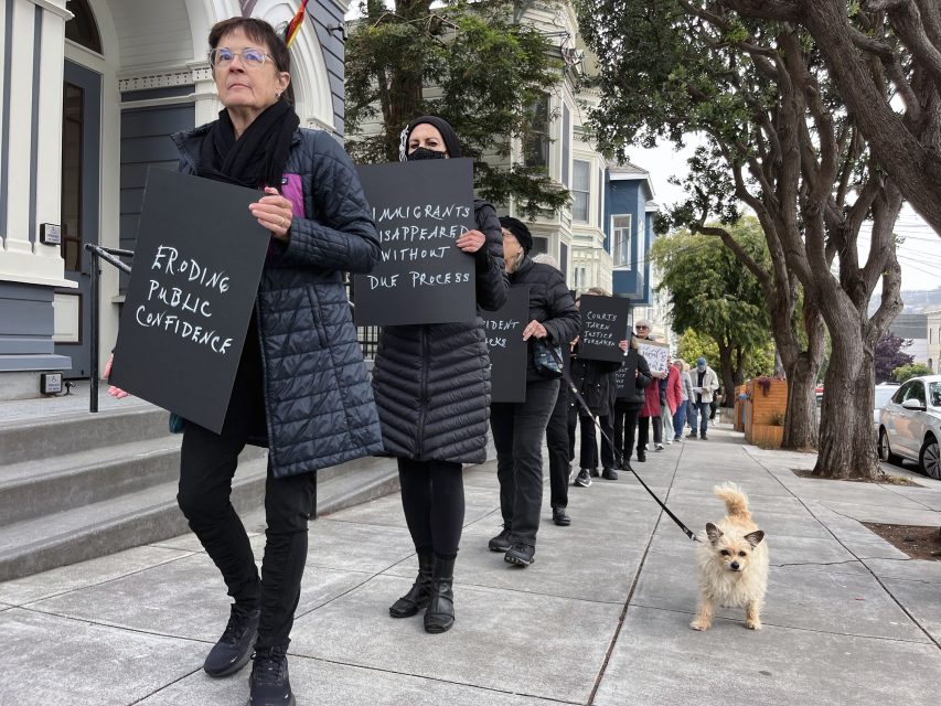 A group of people in dark coats walk in a line on a sidewalk holding black protest signs; a small dog on a leash walks beside them.