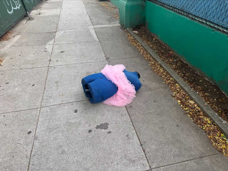 A blue dress form lying on a sidewalk, partly covered with a pink tulle fabric, next to a green wall and metal fence.