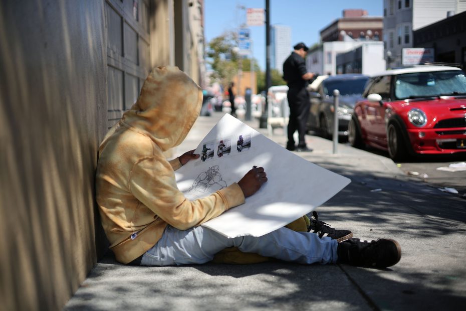 A person in a yellow hoodie sits on a sidewalk drawing on a large sheet of paper, with a street scene and parked cars in the background.