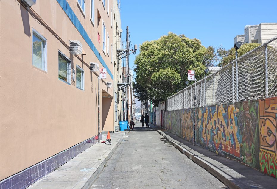 A narrow city alleyway with graffiti-covered walls, a beige building on the left, and a few people walking in the distance on a sunny day.