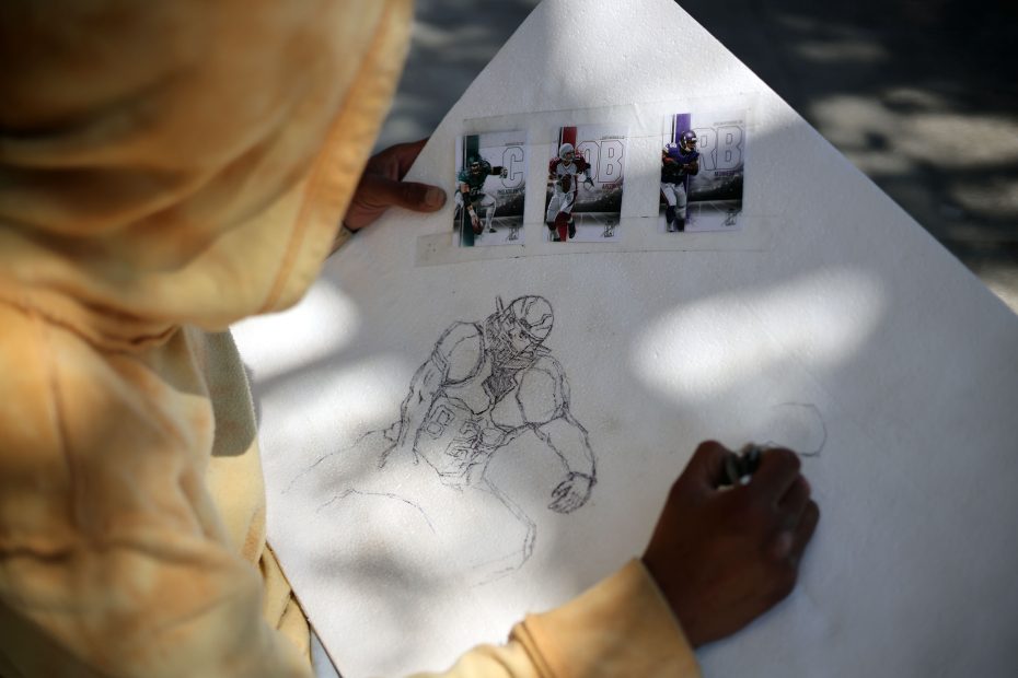 A person in a yellow hoodie sketches an American football player on a white board, referencing three printed photos of football players attached above the drawing.