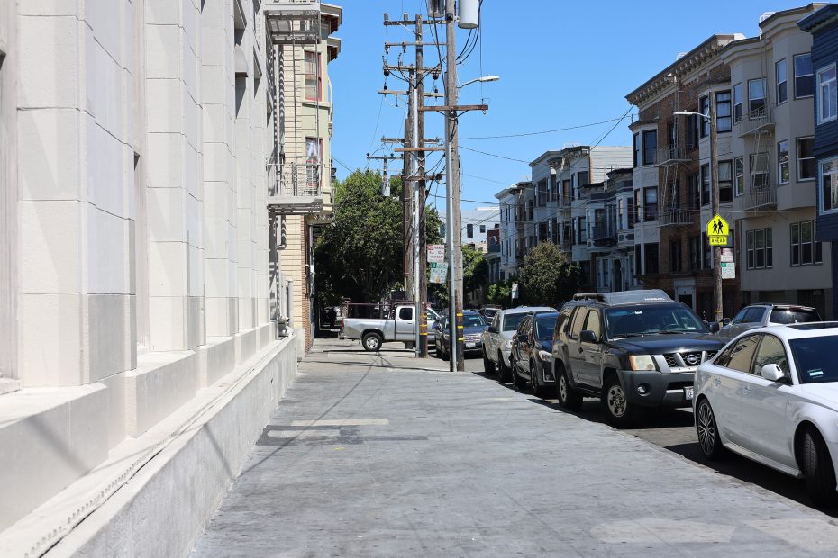 A city street with parked cars, apartment buildings, and power lines on a sunny day; the sidewalk is empty.