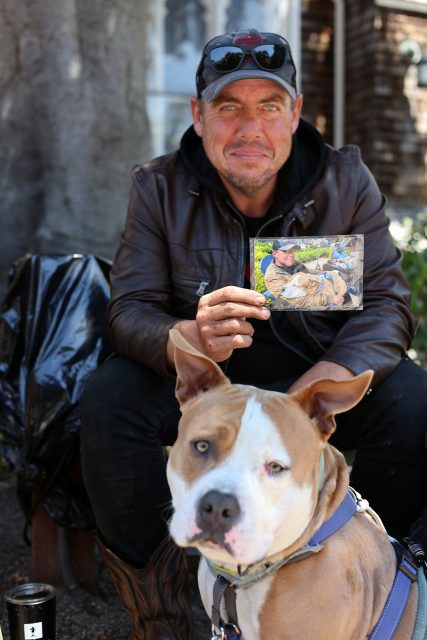 A man in a brown leather jacket holds a photo of himself and his dog while sitting outdoors, with his dog wearing a harness in the foreground.