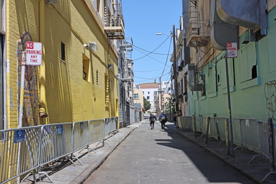 A narrow urban alleyway lined with yellow and green buildings, metal barricades, and "No Parking" signs; two people walk in the distance under a clear sky.