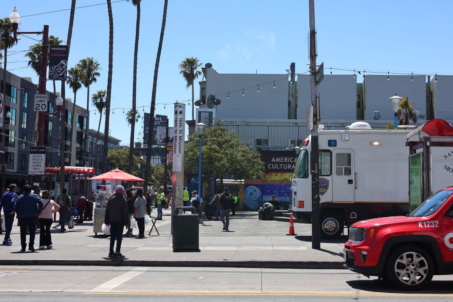 Street scene with people walking, food stands, and emergency vehicles parked near a modern building with palm trees and a sign reading "AMERICAN CULTURAL.