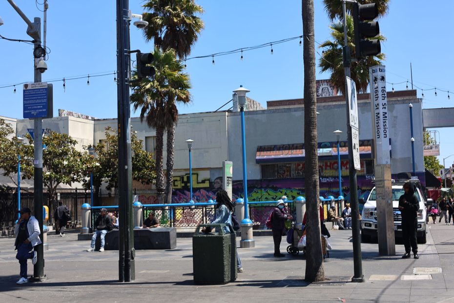 People stand and walk around an urban plaza with palm trees, streetlights, and a graffiti-covered building in the background on a sunny day.