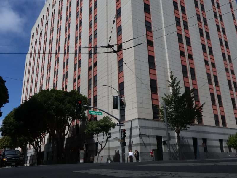 Tall office building with red and gray exterior, trees along the sidewalk, traffic lights, and street signs at an urban intersection during daytime.