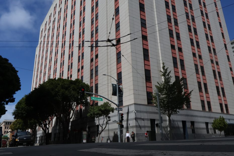 Tall office building with red and gray exterior, trees along the sidewalk, traffic lights, and street signs at an urban intersection during daytime.