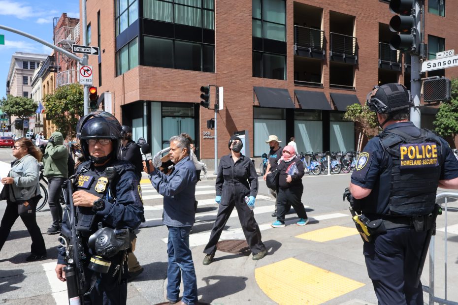 Police officers stand at a city intersection while pedestrians and masked individuals cross the street during daytime.