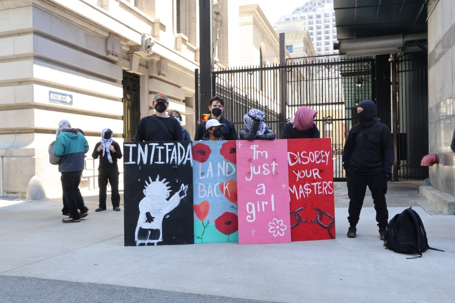 A group of masked protesters stands on a street holding painted signs with slogans including "INTIFADA," "LAND BACK," "I'm just a girl," and "DISOBEY YOUR MASTERS.