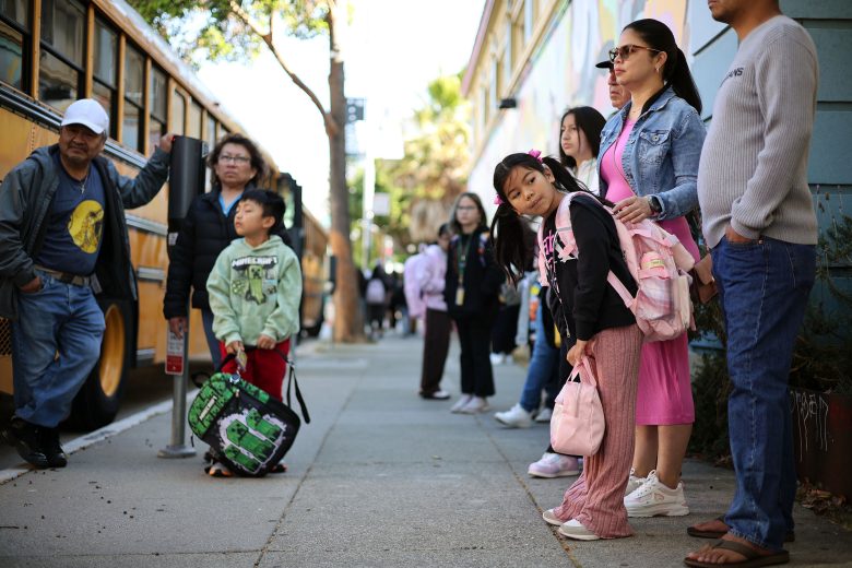 A group of adults and children with backpacks wait on a sidewalk near a yellow school bus and a building.