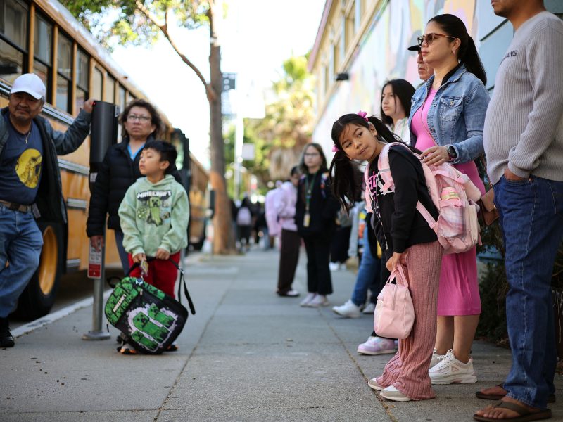 A group of adults and children with backpacks wait on a sidewalk near a yellow school bus and a building.