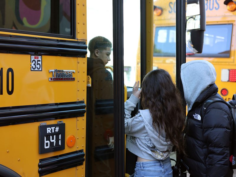 Two students wait to board a yellow school bus while another student steps inside through the open door.