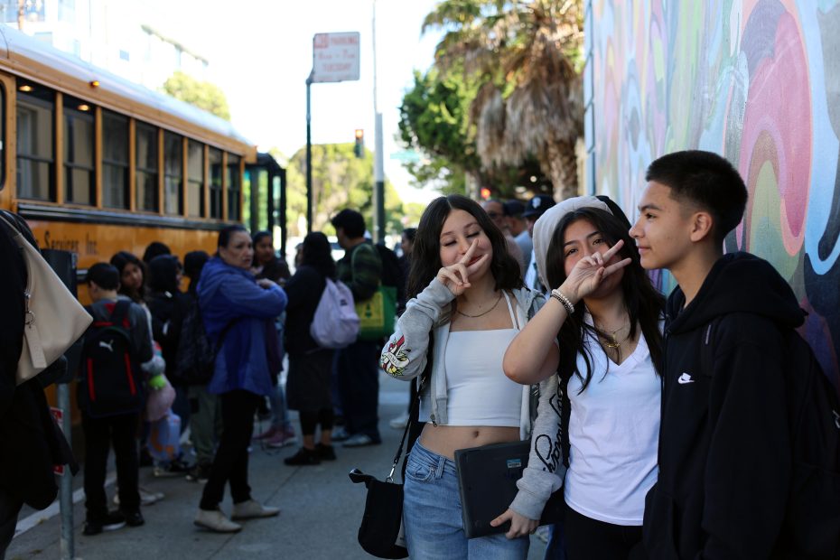 Three teens pose for a photo, two making peace signs, in front of a mural. Behind them, people wait near a yellow school bus on a city street.