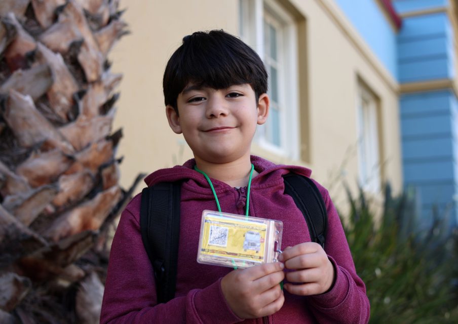A boy in a maroon hoodie and backpack stands outside, holding up a yellow ID badge on a green lanyard and smiling at the camera.