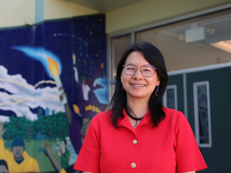A woman in a red shirt stands smiling in front of a building with a colorful mural featuring various figures and a rocket launch.
