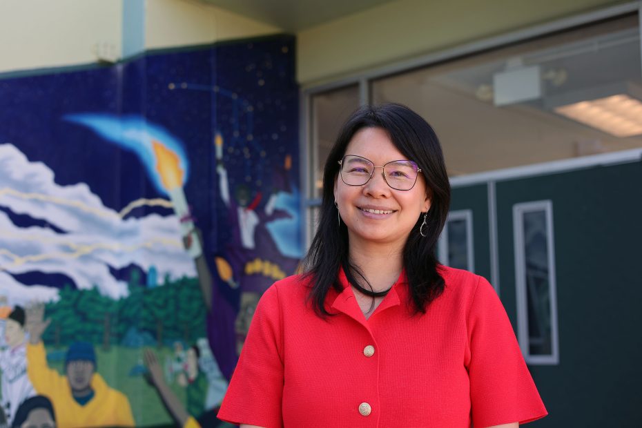 A woman in a red shirt stands smiling in front of a building with a colorful mural featuring various figures and a rocket launch.
