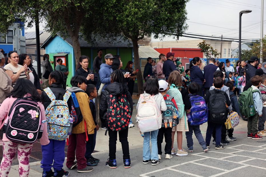 A group of children wearing backpacks stand in a line outdoors, with adults gathered nearby at what appears to be a school.
