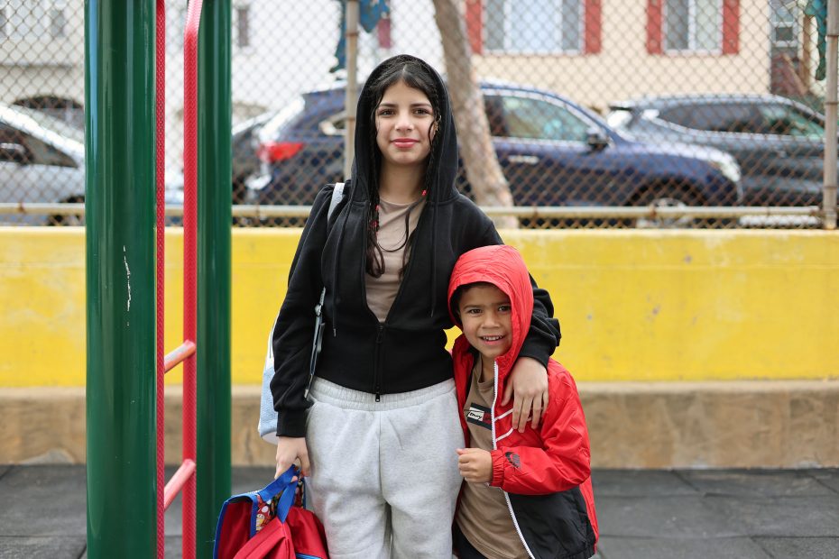 Two children stand together outdoors by a playground, with the older child holding a bag and the younger child wearing a red hooded jacket and smiling.